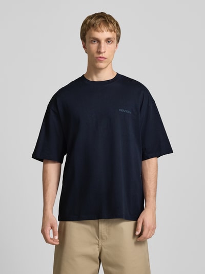 Männermodel trägt Oversized T-Shirt mit Logo von REVIEW in Schwarz, reine Baumwolle, lockerer Schnitt, Rundhalsausschnitt, Ganzkörperansicht von vorne.