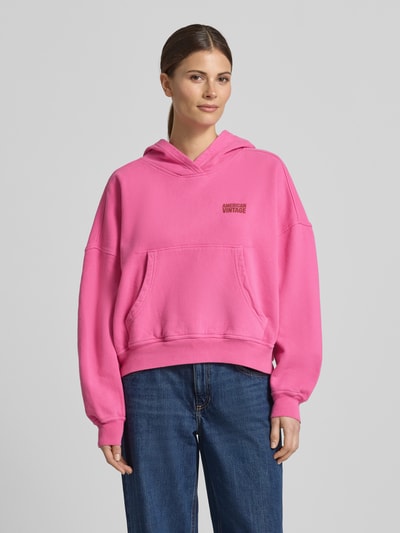 Kobiece model prezentuje bluzę z kapturem z kieszenią kangurka Hoodie mit Känguru-Tasche Modell 'PLIZZY' od American Vintage z bawełny, krój relaxed fit, logo, kolor różowy, pełny przód, zdjęcie całej sylwetki z przodu.