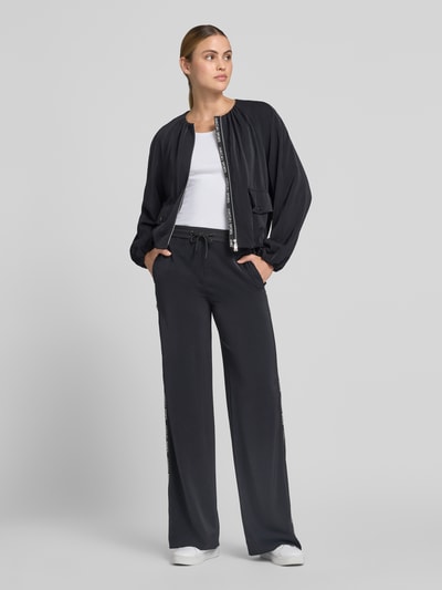 Frauenmodel trägt Schlupfhose mit seitlichen Label-Details und elastischem Bund von Marc Cain in Schwarz, Seitliche Eingrifftaschen, Relaxed-Fit, Tunnelzug, Bundweite bei Größe 36: 39 cm, Innenbeinlänge bei Größe 36: 85 cm, Ganzkörperansicht von vorne.