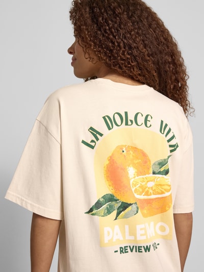 Frauenmodel trägt T-Shirt mit rückseitigem Motiv-Print von REVIEW in Creme mit gelbem und grünem Motiv, Oversized, Reine Baumwolle, Rückenlänge 68 cm, Ganzkörperansicht Rückansicht.