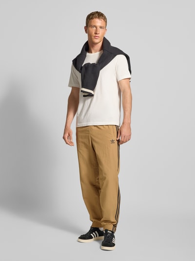 Męski model nosi Sweatpants mit elastischem Bund od adidas Originals w kolorze beżowym z czarnymi paskami, luźny krój, całkowity widok z przodu.