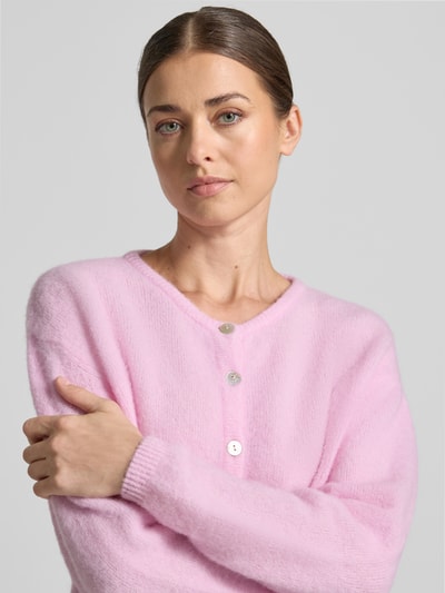 Frauenmodel trägt Strickjacke mit Woll-Anteil Modell 'VITOW' von American Vintage in Rosa, lockere Passform, Nahaufnahme Oberkörper. Elastischer Material-Mix, Rundhalsausschnitt, Knopfleiste, gerippte Abschlüsse.