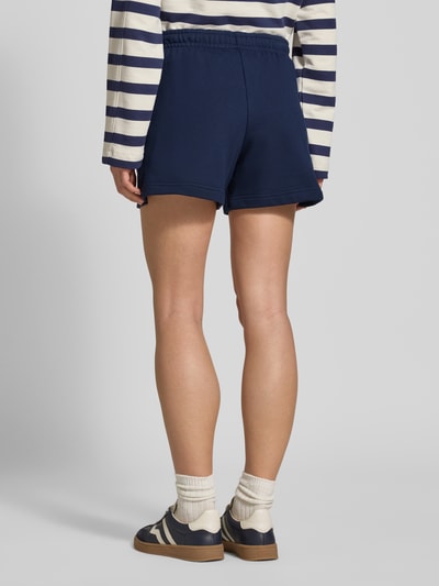 Modelka nosi Regular Fit Sweatshorts aus reiner Baumwolle von Lacoste w jednolitym kolorze, z wyhaftowanym logo, elastycznym ściągaczem, wpuszczanymi kieszeniami bocznymi oraz tunelem; krój regular fit z czystej bawełny, widok całej sylwetki od tyłu.