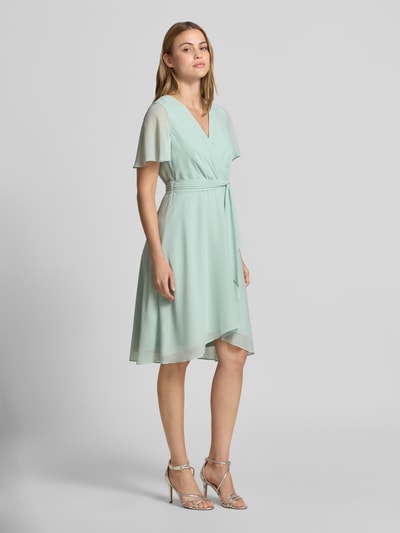 Frauenmodel trägt Knielanges Cocktailkleid in Wickel-Optik von Christian Berg Cocktail aus Chiffon mit A-Linie, V-Ausschnitt, Bindegürtel, Knielänge in hellem Pastellgrün, fließende Textur, feine Struktur, Ganzkörperansicht von vorne. Brustweite 33 cm, Rückenlänge 100 cm, Ärmellänge 22 cm bei Größe 36.