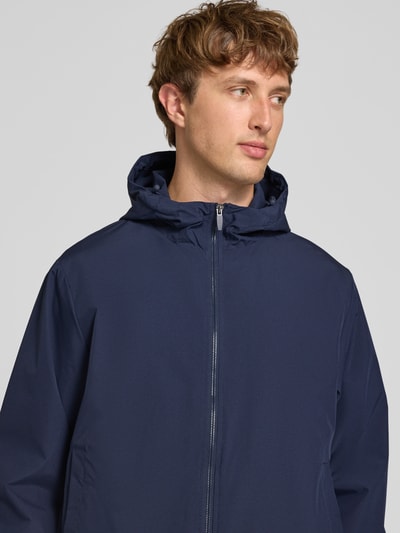 Männermodel trägt Regular Fit Jacke mit Kapuze Modell 'Charge' von JACK & JONES in Dunkelblau, glatte Textur, Nahaufnahme Oberkörper. Rückenlänge bei Größe M: 68 cm, Schulterbreite: 50 cm, Brustweite: 60 cm, Ärmellänge: 66 cm, Reißverschluss.