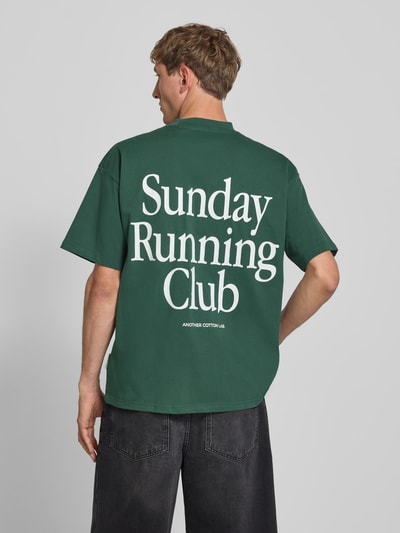 Männermodel trägt T-Shirt mit Print und Rundhalsausschnitt von ANOTHER COTTON LAB, męski t-shirt, bawełna, krój relaxed fit, nadruk, okrągły dekolt, długość tyłu w rozmiarze M: 69 cm, zielony, prostokątne formy, miękka faktura, Ganzkörperansicht Rückansicht.