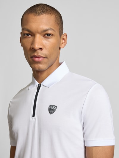 Mężczyznamodel nosi Slim Fit Poloshirt aus Viskose-Mix od EA7 Emporio Armani, biała koszulka z detalem z logo, kołnierzykiem polo, elastyczne zakończenia rękawów, elastyczna mieszanka wiskozy, krój slim fit, długość tyłu w rozmiarze M: 60 cm, zbliżenie na górną część ciała.