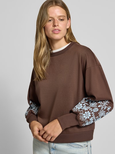 Frauenmodel trägt Relaxed Fit Sweatshirt aus reiner Baumwoll-Mix Modell 'BROOKE' von ONLY, braun mit blauen Blumenärmeln, Nahaufnahme Oberkörper, Damen Sweatshirt von ONLY