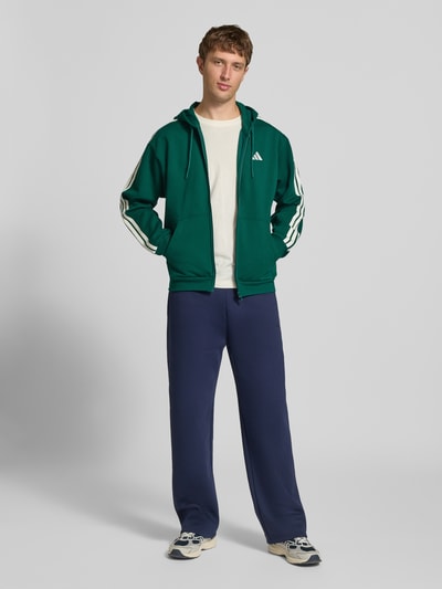 Männermodel trägt Regular Fit Sweatjacke aus Baumwoll-Mix von adidas Sportswear in Dunkelgrün mit weißen Details, Ganzkörperansicht von vorne.
