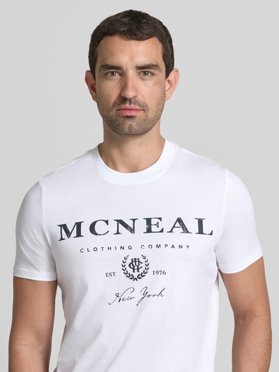 Męski model nosi T-Shirt mit Label-Print und Rundhalsausschnitt od MCNEAL z czystej bawełny, krój regular fit, nadruk z logo, okrągły dekolt, długość tyłu w rozmiarze M: 68 cm, biały, czarne napisy, gładka faktura, zbliżenie na górną część ciała