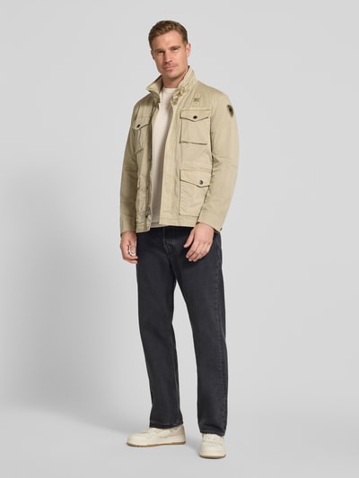 Mężczyznа nosi kurtkę Jacke mit Stehkragen und Pattentaschen Modell 'Ashmont' von Blauer Usa z mieszanki bawełny i elastanu, krój regular fit, kolor beżowy, cztery kieszenie z patką i stójka, długość tyłu w rozmiarze M: 72 cm, szerokość ramion w rozmiarze M: 40 cm, szerokość klatki piersiowej w rozmiarze M: 52 cm, długość rękawów w rozmiarze M: 63 cm, całkowity widok z przodu