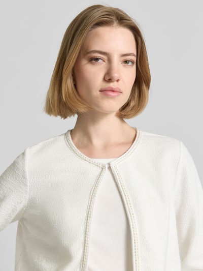 Kobiece model nosi Regular Fit Jacke mit Zierbordüre od More & More, elastyczna mieszanka materiałów z mieszanką bawełny, otwarty krój, ozdobna lamówka, biała faktura, długość tyłu 48 cm, długość rękawów 47 cm, zbliżenie na górną część ciała.