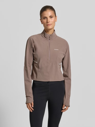 Frauenmodel trägt Cropped Pullover für intensive Anstrengungen und Beschleunigung von Salomon in Taupe mit Stehkragen, Reißverschluss, Logo, Material-Mix mit Stretch-Anteil, Rückenlänge bei Größe S: 47 cm, Ganzkörperansicht von vorne.