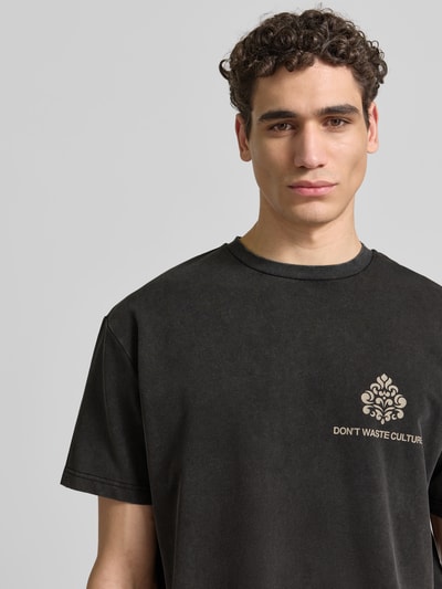 Männermodel trägt Relaxed Fit T-Shirt aus Baumwoll-Mix von Don’t Waste Culture in Schwarz mit Rundhalsausschnitt, Used-Look, Label-Details, Nahaufnahme Oberkörper. Rückenlänge bei Größe M: 68 cm.