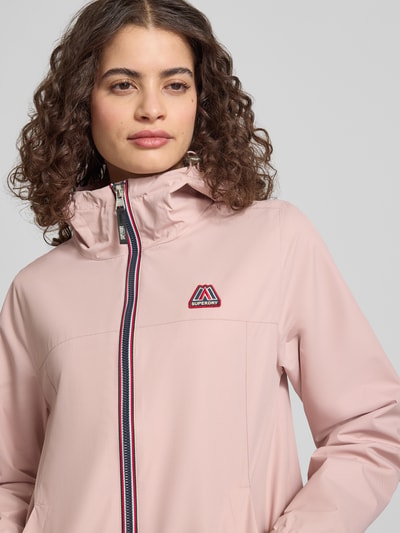 Modelka nosi Regular Fit Windbreaker mit Logo-Applikation od Superdry, kolor jasnoróżowy, kaptur ze ściągaczem, krój regular fit, kieszenie boczne, zamek z przodu, detal z logo, długość pleców w rozmiarze S: 25 cm, szerokość ramion w rozmiarze S: 44 cm, szerokość klatki piersiowej w rozmiarze S: 49 cm, długość rękawa w rozmiarze S: 65 cm, zbliżenie na górną część ciała