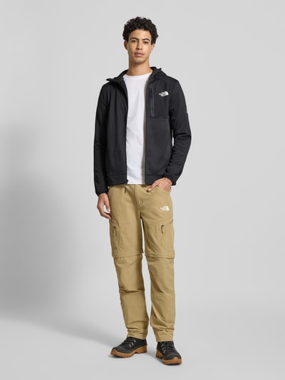 Männermodel trägt schwarze Sweatjacke mit Napoleontasche Modell 'MOUNTAIN ATHLETICS' von The North Face, Körpernahe Passform, Kapuze, Label-Details, durchgehender Reißverschluss, elastische Abschlüsse, Ganzkörperansicht von vorne.