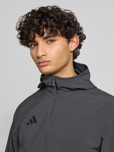 Männermodel trägt Slim Fit Funktionsjacke aus schnelltrocknendem Material von adidas Sportswear, efekt: jednolity kolor, krój slim fit, kaptur, zamek błyskawiczny, nadruk z logo, paski w kontrastowym kolorze, długi rękaw, szybkoschnący, materiał pochłaniający pot, kieszenie zapinane na zamek błyskawiczny; Nahaufnahme Oberkörper