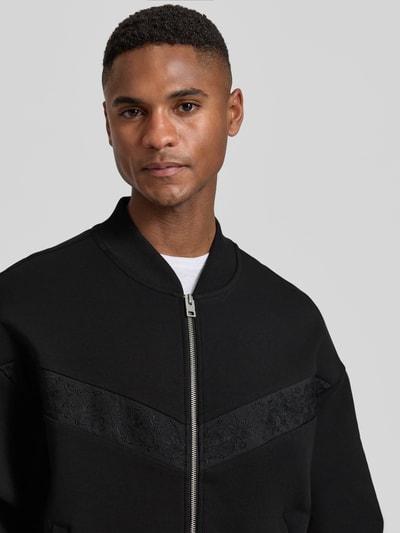 Männermodel trägt Oversized Sweatjacke aus Baumwoll-Mix Modell 'DROMZIP' von HUGO in Schwarz, Logo-Patch, Nahaufnahme Oberkörper.