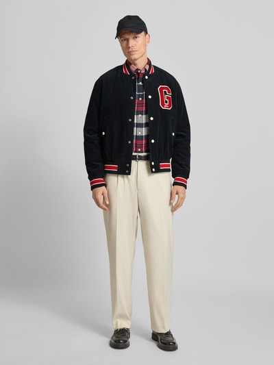 Männermodel trägt College-Jacke aus Cord mit Label-Detail von Gant, Regular-Fit, schwarze und rote Farbakzente, Reine Baumwolle, Stehkragen, Knopfleiste, Eingrifftaschen, Rückenlänge 66 cm, Schulterbreite 46 cm, Brustweite 53 cm, Ärmellänge 67 cm, Ganzkörperansicht von vorne.
