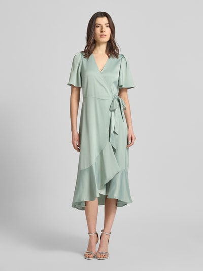 Frauenmodel trägt Cocktailkleid mit Volantsaum Modell 'THEA' von YAS, Satin, Taillierter Schnitt, Midikleid, Wickel-Optik, V-Ausschnitt, Schmetterlingsärmel, fließender Stoff in sanfter Pastellfarbe, Ganzkörperansicht von vorne.