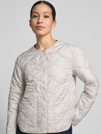Vrouwenmodel draagt Steppjacke mit Rundhalsausschnitt van camel active in lichte crèmekleur, rechte pasvorm, stikseldetails, drukknopen, vooraanzicht buste close-up, ruglengte 60 cm, schouderbreedte 43 cm, borstwijdte 53 cm, mouwlengte 64 cm.