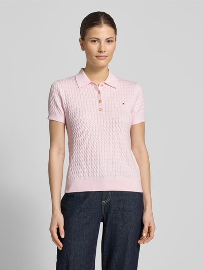 Frauenmodel trägt Slim Fit Poloshirt aus reiner Baumwolle von Tommy Hilfiger, rosa, Zopfmuster, V-Ausschnitt, Logo-Detail, Ganzkörperansicht von vorne.