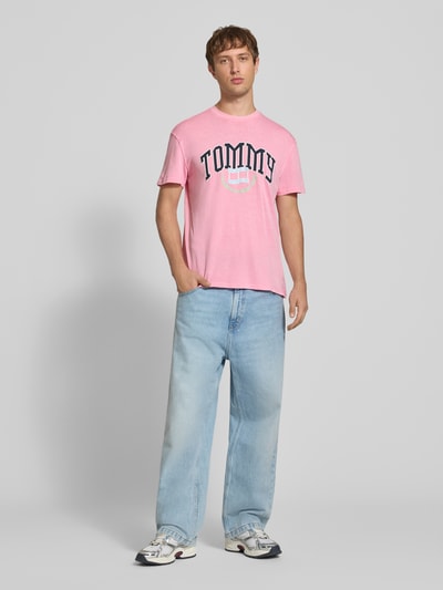 Männermodel trägt Regular Fit T-Shirt aus reiner Baumwolle von Tommy Jeans in Rosa mit Print und heller Jeans, Ganzkörperansicht von vorne.