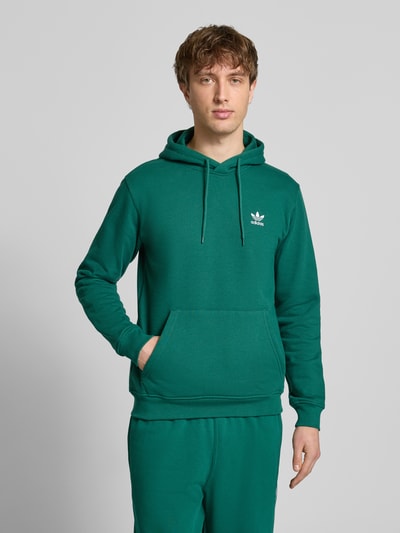 Männermodel trägt Hoodie mit Kapuze von adidas Originals in Grün mit Känguru-Tasche und Label-Stitching, Nahaufnahme Oberkörper.