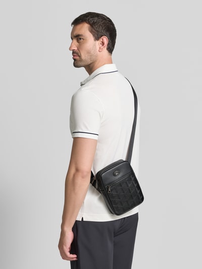 Männermodel trägt schwarze Umhängetasche mit abnehmbarem Schulterriemen von Lacoste, dezente Oberflächenstruktur, Ganzkörperansicht Rückansicht. Herren Umhängetasche von Lacoste.