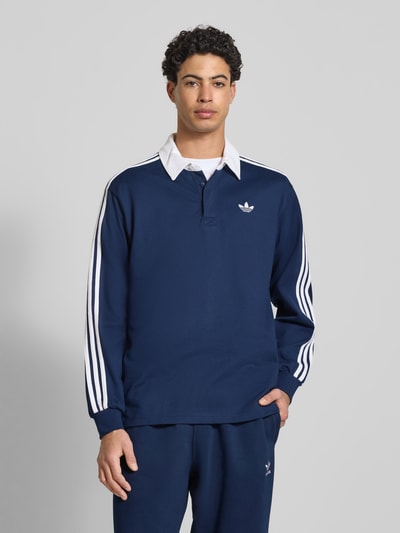 Männermodel trägt dunkelblaues Poloshirt mit Label-Stitching von adidas Originals, reine Baumwolle, Regular Fit, Ganzkörperansicht von vorne.