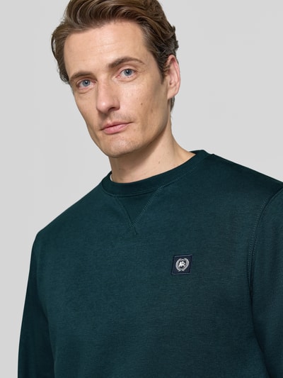 Männermodel trägt Regular Fit Sweatshirt aus Baumwoll-Mix von LERROS in Dunkelgrün mit Logo-Patch und gerippten Abschlüssen, Nahaufnahme Oberkörper.