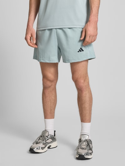 Männermodel trägt Regular Fit Shorts mit Logo-Print von adidas Training in Hellblau, elastischer Bund, Reißverschlusstaschen, Nahaufnahme Oberschenkel.
