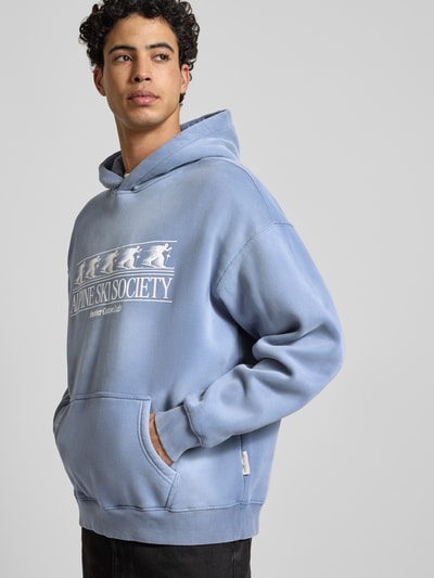 Männermodel trägt Oversized Hoodie mit Kapuze von ANOTHER COTTON LAB in Hellblau, Baumwoll-Mix, Label-Print, Känguru-Tasche, Ganzkörperansicht von vorne.