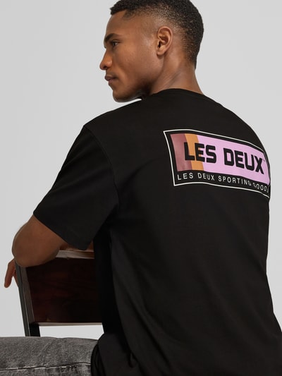 Męski model nosi T-Shirt mit Logo-Print Modell 'Chase' von Les Deux, czarna bawełna, prosty krój, logo na plecach, ujęcie z tyłu