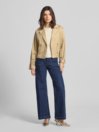 Frauenmodel trägt Cropped Trenchcoat mit Reverskragen Modell 'Fatou' von Guess aus Baumwoll-Mix, taillierte Passform, Doppelknopfleiste, Stoffgürtel in Beige, Ganzkörperansicht von vorne.