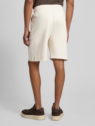 Męski model nosi szorty z wpuszczanymi kieszeniami model 'Joseignacio' od JOOP! Collection, mieszanka bawełny z dodatkiem streczu, krój loose fit, elastyczny pas z trokiem, naszywka z logo, kolor jasny, szerokość w rozmiarze M: 42 cm, długość wewnętrzna nogawki w rozmiarze M: 22 cm, widok całej sylwetki z tyłu.