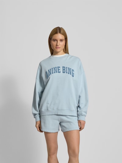 Frauenmodel trägt Oversized Sweatshirt mit Logo-Detail von Anine Bing in Hellblau, Reine Baumwolle, Oversized-Fit, Rundhalsausschnitt, elastische Abschlüsse, Damen Sweatshirt, Rückenlänge bei Größe S 65 cm, Ganzkörperansicht von vorne.