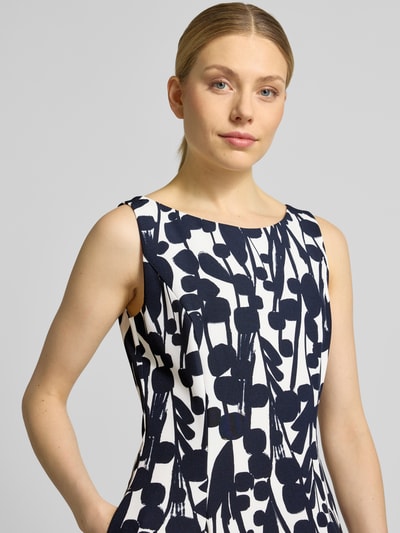 Vrouwenmodel draagt knielanges cocktailjurk met naadritssluiting van Vera Mont, elastische materiaalmix, plooien, korte mouw, asymmetrische V-hals, eng aansluitende pasvorm, wit met donkerblauwe print, dames, detailopname bovenlichaam.