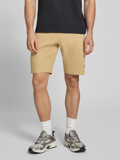 Männermodel trägt Straight Fit Shorts mit Logo-Stitching Modell 'Fillip' van Blend in beige structuurpatroon, puur katoen, steekzakken, achterzakken, elastische band met tunnelkoord, bandwijdte maat M 39 cm, binnenbeenlengte maat M 26 cm. Ganzkörperansicht von vorne.