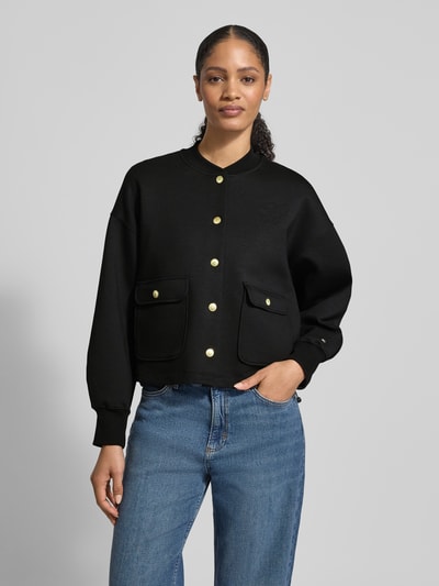 Frauenmodel trägt Regular Fit Bomberjacke aus Baumwoll-Mix Modell 'GOLD BUTTON BOMBER' von Tommy Hilfiger in Schwarz mit goldenen Knöpfen, unifarben, Logo-Stitching, College-Kragen, Pattentaschen, gerippte Ärmelabschlüsse, Tunnelzug, Ganzkörperansicht von vorne.