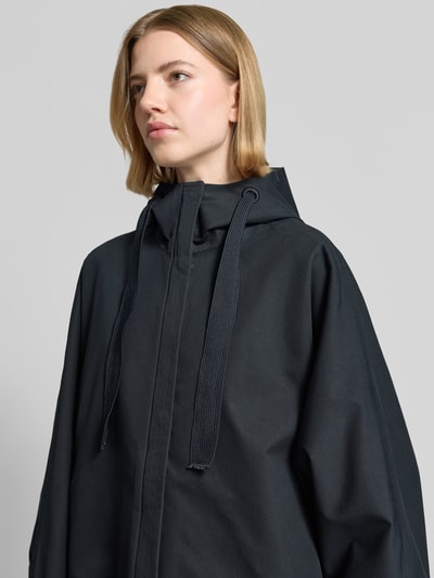 Modelka nosi kurtkę Jacke mit Kapuze und Eingrifftaschen Modell 'FEEL' od G-LAB, mieszanka bawełny, luźny krój, kaptur z trokiem, wpuszczane kieszenie, patki przy mankietach na zatrzaski, długość tyłu w rozmiarze S 78 cm, szerokość ramion 49 cm, szerokość klatki piersiowej 84 cm, długość rękawów 58 cm, kolor ciemny, gładka tekstura, zdjęcie zbliżenie górnej części ciała z przodu.
