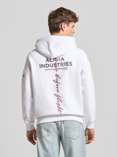 Männermodel trägt Hoodie mit Label-Details von Alpha Industries, weiß, Baumwoll-Mix, Kapuze, Reißverschlusstasche, Lockerer-Schnitt, Ganzkörperansicht Rückansicht.