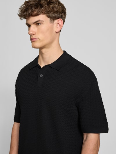 Männermodel trägt Relaxed Fit Poloshirt Modell 'FERDINAND' von Redefined Rebel, schwarze Reine Baumwolle, feines Strickmuster, Knopfleiste, Nahaufnahme Oberkörper. Rückenlänge bei Größe M: 70 cm.