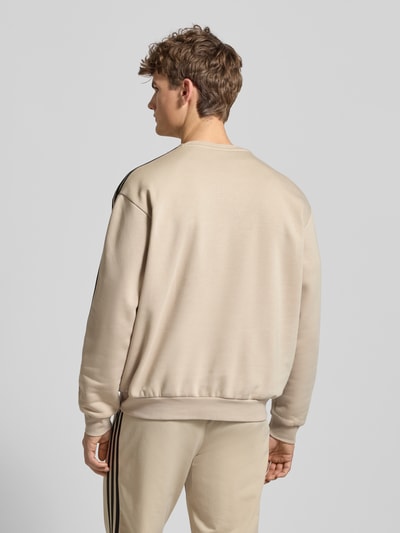 Męski model nosi Sweatshirt mit Label-Stitching od adidas Originals, mieszanka bawełny, krój regular fit, prążkowane wykończenia, okrągły dekolt, długość tyłu 69 cm, długość rękawów 60 cm, jasny beżowy kolor, gładka tekstura, całkowity widok z tyłu.