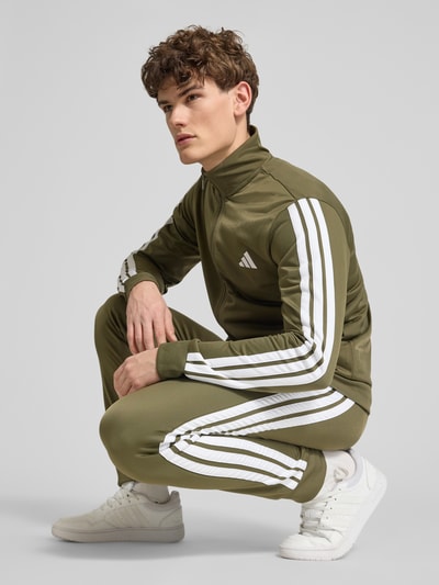 Männermodel trägt Regular Fit Sweat-Anzug mit Kontraststreifen von adidas Sportswear in Oliv mit Optik: Unifarben, Ganzkörperansicht von vorne.