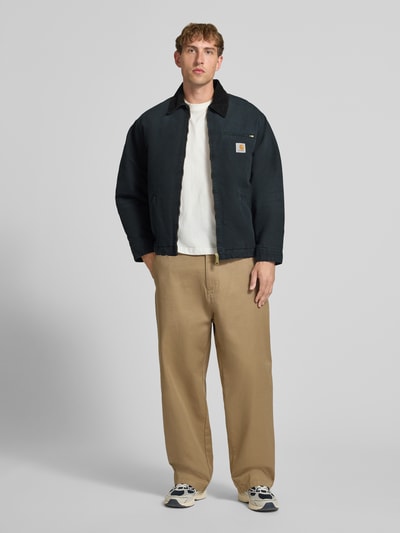 Männermodel trägt Blouson mit Brusttasche Modell 'DETROIT' von Carhartt Work In Progress, dunkle Farben, Cord-Kragen, Reißverschlüsse, Ganzkörperansicht von vorne.