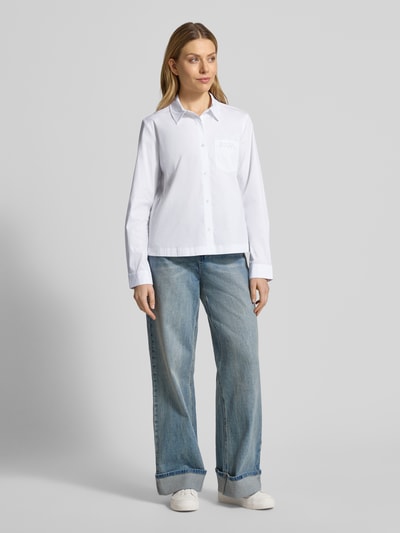 Modelka kobieca nosi Regular Fit Hemdbluse mit Brusttasche Modell 'Bella' od JOOP!, jasnobiały, długi rękaw, kołnierz koszulowy, bawełna elastyczna, guziki, kieszeń z logo, krój regular fit, widok całej sylwetki z przodu.