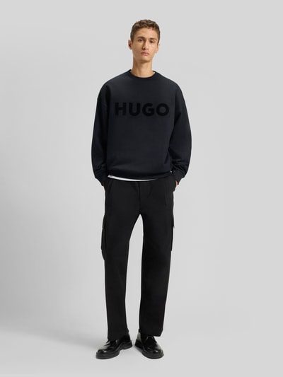Männermodel trägt Oversized Sweatshirt aus reiner Baumwolle Modell 'DINEX' von HUGO in Schwarz mit Rundhals und Label Print, Ganzkörperansicht von vorne.