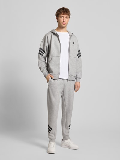 Männermodel trägt Sweatjacke mit Kapuze und Logo von adidas Sportswear im Relaxed-Fit, Material-Mix mit Baumwoll-Anteil, grau mit schwarzen Details, Känguru-Tasche, Reißverschluss, Logo, Ganzkörperansicht von vorne. Rückenlänge 68 cm, Ärmellänge 70 cm, Größe M.