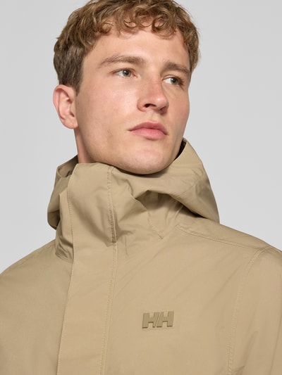 Männermodel trägt Regular Fit Mantel mit Kapuze Modell 'Brussel' von Helly Hansen, beige, Logo-Detail, Nahaufnahme Oberkörper. Rückenlänge bei Größe M: 96 cm, Schulterbreite: 46 cm, Brustweite: 51 cm, Ärmellänge: 66 cm.
